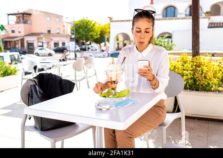 Belle femme moderne et élégante assise dans un café bar déhore table appréciant sa grande glace aromatisée aux fruits à l'aide de mobile téléphone avec connexion internet Banque D'Images