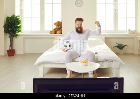 Drôle de barbu gros homme actif fan de football regarder un match à la télévision à la maison assis sur un canapé. Banque D'Images