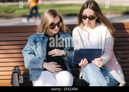 Femme enceinte avec sa meilleure amie assise dans le parc et à la recherche de poussette de bébé à l'aide d'un comprimé. Deux femelles en lunettes de soleil choisissent des vêtements pour bébé Banque D'Images