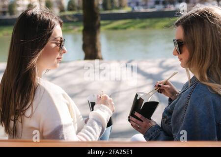 Deux jolie femme mangeant fastfood ouddood. Belle fille assise sur le banc dans le parc et de prendre la nourriture asiatique. Concept de plats à emporter Banque D'Images