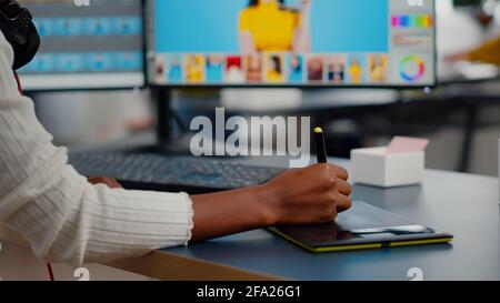 Gros plan de retouche photo africaine tenant un stylet retouche photo de client à l'aide d'un ordinateur avec deux écrans. Éditeur graphique retouche des ressources de dessin sur tablette graphique dans un bureau d'agence de création Banque D'Images