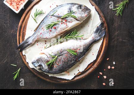 poisson dorado frais cru sur une planche à découper en bois avec du sel de l'himalaya rose, des grains de poivre rose et du romarin Banque D'Images