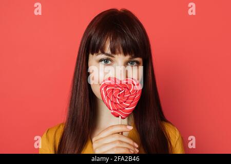 Gros plan en studio de charmante jeune femme aux cheveux rouges, portant une chemise jaune, tenant un lollipop rouge en forme de coeur, couvrant son nez et ses lèvres, posant sur un fond rouge. Banque D'Images