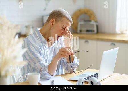 Une femme d'âge moyen, fatiguée et endormie, massante le nez et bâillante tout en travaillant sur un ordinateur portable en ligne depuis la maison, assise dans la cuisine moderne à la maison. Concept de personnes âgées et de travail à distance Banque D'Images
