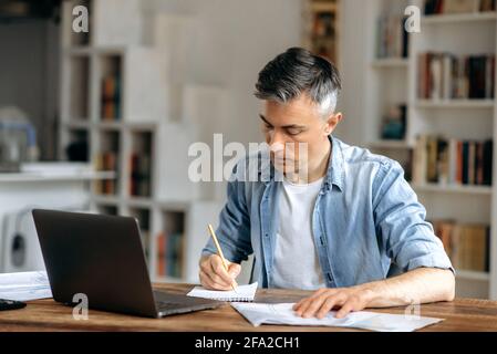 Manager, coach ou freelance à cheveux gris, élégant et concentré, habillé dans des vêtements décontractés et élégants, travaillant à la maison avec un ordinateur portable et des documents à la table, préparant un projet d'entreprise, prenant des notes Banque D'Images