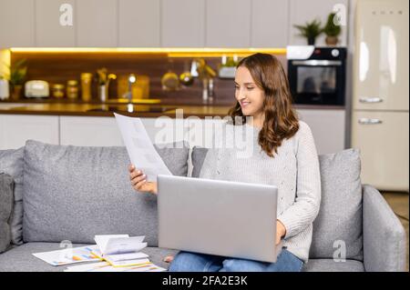 Femme pigiste gaie utilisant un ordinateur portable pour travailler à distance depuis la maison, femme assise sur le canapé et regarde les feuilles de documents de travail, prépare le rapport, travaille avec le projet depuis la maison Banque D'Images