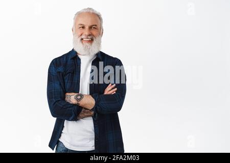 Vieux homme plein d'assurance souriant et riant. Garçon de taille basse barbu senior avec tatouages croiser les bras sur la poitrine, regardant déterminé et heureux à la caméra Banque D'Images