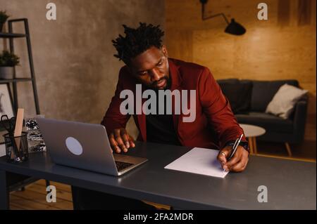Beau Noir Africain américain homme travaillant sur ordinateur portable tout en étant assis derrière le bureau dans le salon confortable. Travailleur indépendant à domicile Banque D'Images