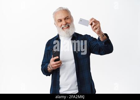 Homme senior heureux tenant le smartphone, levant la main et montrant la carte de crédit avec le sourire satisfait, hocher dans l'approbation, recommander le paiement en ligne, mobile Banque D'Images