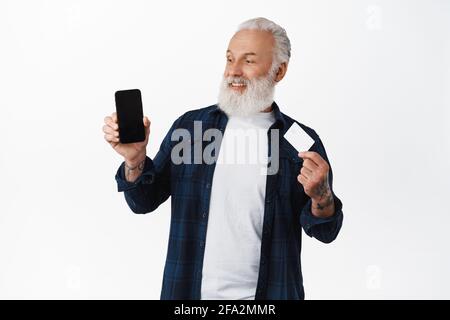 Homme senior heureux tenant la carte de crédit de banque, regardant satisfait à l'écran vide de téléphone mobile avec votre application ou page Web d'achat, debout contre blanc Banque D'Images