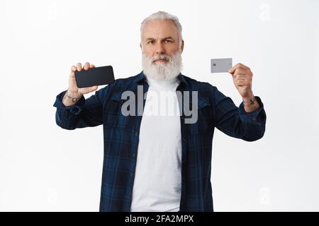 Homme barbu sérieux et mûr montrant l'écran du téléphone portable et la carte de crédit, montrant quelque chose sur l'écran du smartphone, debout sur fond blanc Banque D'Images