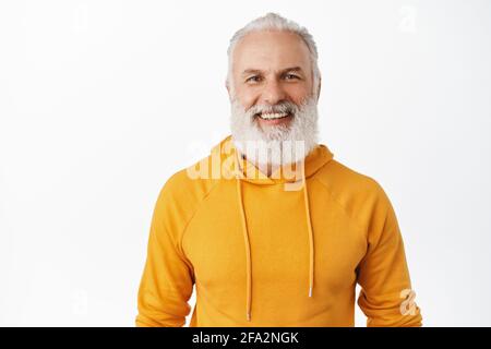 Gros plan sur un vieil homme charmant avec une barbe, souriant et plein de gaieté, debout dans un élégant sweat à capuche orange sur fond blanc Banque D'Images