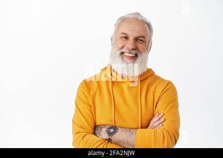 Beau vieil homme avec des tatouages riant et souriant, montrant des émotions authentiques, des bras croisés sur la poitrine, se tenant détendu sur fond blanc Banque D'Images