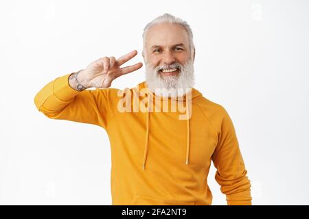 Positif hipster senior avec la barbe et les tatouages montre le V-signe de peave près de l'oeil, souriant et riant heureux, portant le sweat à capuche orange élégant, se tenant dessus Banque D'Images