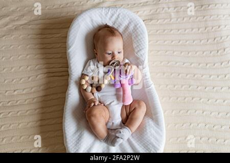 Mignon bébé blond de trois mois couché dans un cocon à la maison. Enfant tient un jouet sonore bourré, suce le mamelon. Banque D'Images