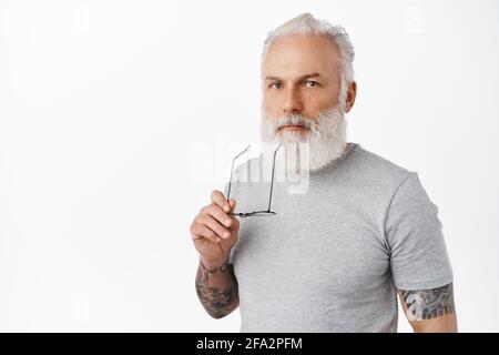 Un vieil homme élégant et attentionné qui touche les lèvres avec un temple de lunettes, qui regarde sérieusement l'appareil photo, pense, réfléchit à la décision, se tient dans un t gris décontracté Banque D'Images