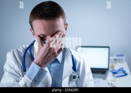 Jeune médecin européen allemand regardant l'appareil photo portant des lunettes et un masque dans le bureau de la clinique sur le lieu de travail avec le regard fatigué. Le sujet est le dur travail de Banque D'Images