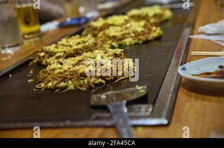 Mise au point sélective sur la première d'une ligne de plusieurs portions d'okonomiyaki sur une longue table de barbecue à l'intérieur d'un Restaurant japonais Banque D'Images