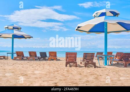 Chaises longues sur la plage. Parasols du soleil avec des sièges sur le lac. La surface de la mer est dégagée sous un ciel bleu. Océan par temps clair. Transats en bois latés. Plage dans une pension ou un hôtel. Vacances. Banque D'Images