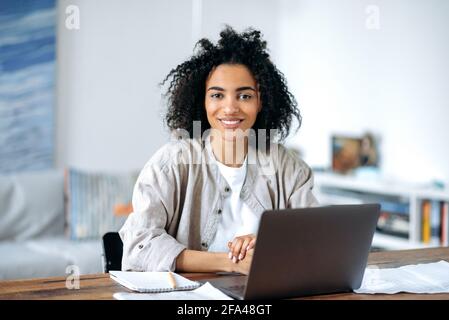 Belle amicale confiante jeune femme afro-américaine dans des vêtements décontractés élégants, indépendant, directeur ou étudiant assis à un ordinateur portable, travaillant ou étudiant à la maison, regardant l'appareil photo, souriant Banque D'Images