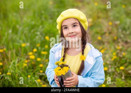 Portrait d'une belle petite fille avec deux queues de porc tient un bouquet de pissenlits jaunes dans une tasse en métal, regarde vers le haut Banque D'Images