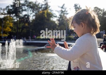 une adorable petite fille regarde la fontaine. elle est petite dans un parc avec des fontaines par temps ensoleillé. week-end en famille. petite fille qui regarde dans une fontaine Banque D'Images