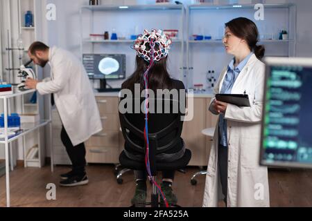 Vue arrière d'une femme portant un casque pour l'acquisition du cerveau en laboratoire de neurologie, neuro science. Système nerveux analysé par une équipe de scientifiques. Scientifique médical regardant à travers le microscope en arrière-plan de développement de traitement. Banque D'Images