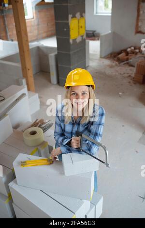 une jolie couche de briques féminine avec un casque de sécurité jaune est scié briques sur un chantier de construction dans la maison et est heureux Banque D'Images