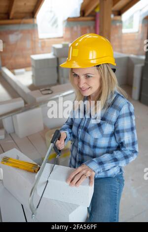 une jolie couche de briques féminine avec un casque de sécurité jaune est scié briques sur un chantier de construction dans la maison et est heureux Banque D'Images