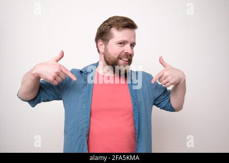 Drôle barbu homme dans des vêtements décontractés constamment se pointe avec les doigts d'index, sourit gentiment et regarde la caméra. Banque D'Images