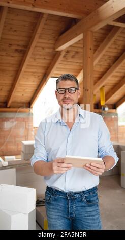 homme architecte d'âge moyen avec des verres noirs se tient sur la construction site dans la maison de loft et tient sa tablette Banque D'Images