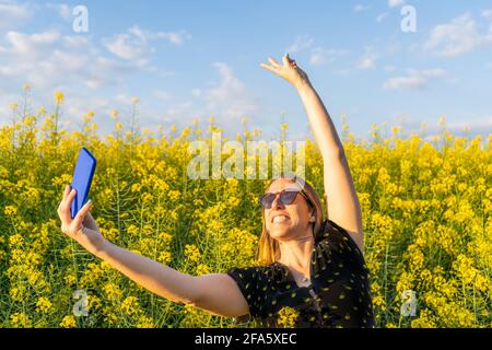 Portrait d'une femme heureuse en lunettes de soleil prenant un selfie et levant le bras dans le champ de fleurs jaunes au coucher du soleil. Banque D'Images