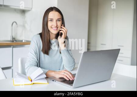 Une jeune femme souriante et positive parle sur un smartphone travaillant avec un ordinateur portable depuis sa maison, une femme tenant une connexion téléphonique avec des clients ou des collègues par téléphone portable regarde la caméra Banque D'Images