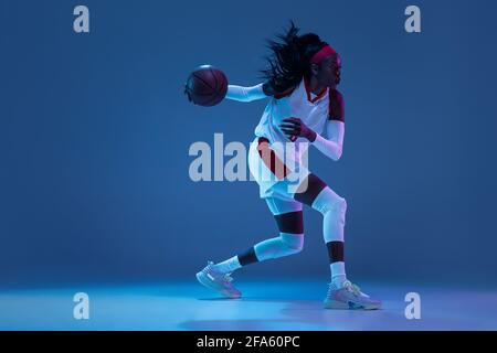 Magnifique joueuse de basket-ball afro-américaine en mouvement et en action avec une lumière néon sur fond bleu. Concept de mode de vie sain, professionnel Banque D'Images