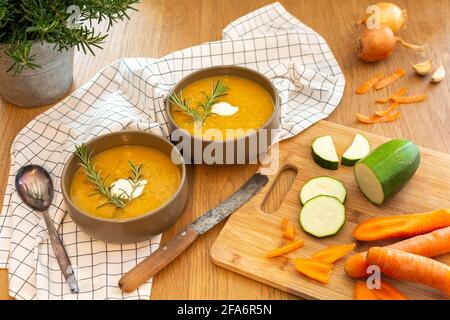 Un bon repas maison contenant une soupe de légumes frais avec des carottes et des courgettes et des oignons servis sur une vieille table en bois avec beaucoup de garniture. Banque D'Images