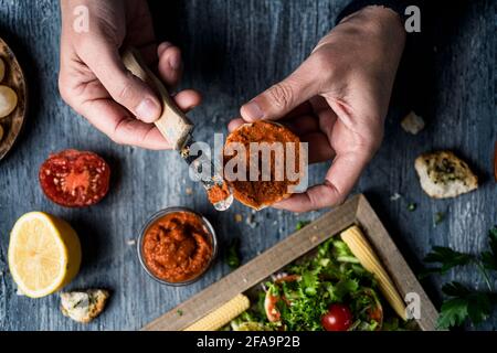 un jeune caucasien répand une pâte végétalienne, faite de tomates séchées et d'amandes, sur un pain grillé de moitié, assis dans un t de bois rustique gris Banque D'Images