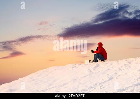 Grimpeur de montagne en manteau rouge vif et lunettes de soleil assis à regarder le lever de soleil orange incroyable sur le sommet de montagne enneigé. Café de haute colline. Banque D'Images