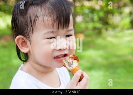 Gros plan adorable petite fille asiatique (thaïlandaise) aimez manger son déjeuner dans le parc, enfant manger du poulet frit sur fond vert flou nature Banque D'Images