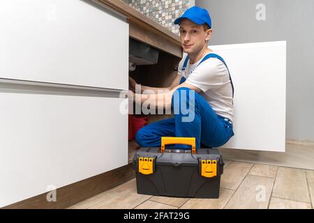 Le handyman en combinaison répare les tuyaux sous l'évier dans la cuisine, à côté d'une boîte d'outils. Regarde la caméra Banque D'Images