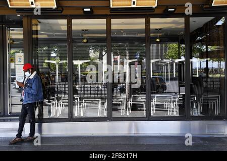 Homme portant une casquette rouge, un masque facial et une veste en jean bleu devant une vitrine fermée avec des chaises empilées à cause du covid à Paris, France Banque D'Images