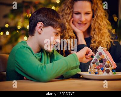 Sourire mignon garçon décorant maison de pain d'épice par la mère pendant Noël Banque D'Images