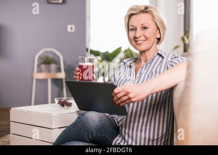Femme souriante tenant une tablette numérique et un verre de jus tout en étant assise à la maison Banque D'Images