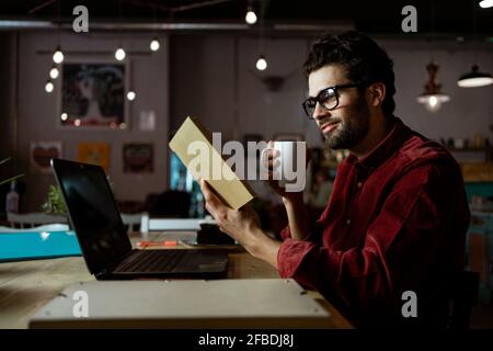 Homme souriant indépendant avec livre de lecture d'ordinateur portable tout en prenant le café dans le café Banque D'Images