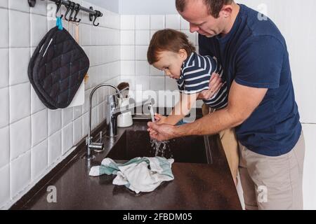 Père transportant son fils tout en se lavant les mains à travers le robinet dans la cuisine à la maison Banque D'Images