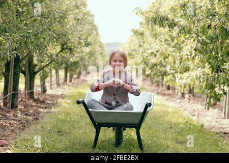Jolie fille en forme de coeur avec les doigts tout en étant assise dans la brouette au verger Banque D'Images