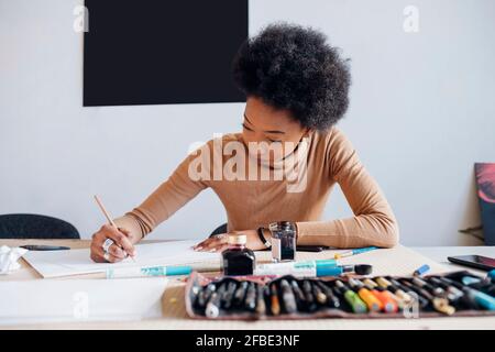 Femme afro écrivant sur papier avec un stylo tout en étant assise table en studio Banque D'Images