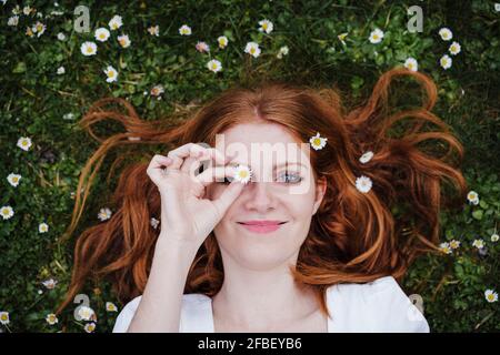 Belle femme souriante couvrant l'œil avec une fleur de pâquerette pendant qu'elle est allongée sur l'herbe Banque D'Images