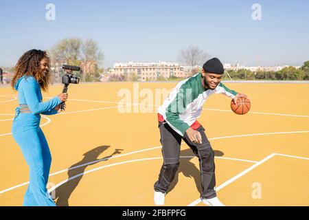 Une femme souriante filmant un ami jouant au basket-ball sur un terrain de sport Banque D'Images