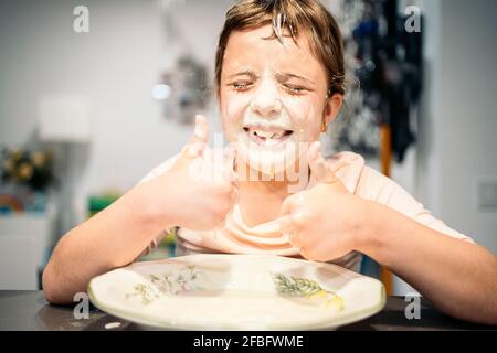 Fille souriante montrant les pouces vers le haut tandis que le visage étalé de farine et de l'eau Banque D'Images