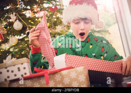 Garçon excité dans chapeau de père Noël déballer cadeau avec arbre de Noël en arrière-plan à la maison Banque D'Images